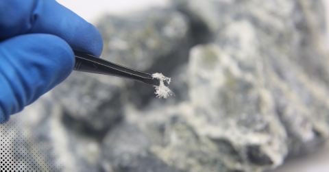 Gloved hand holding tweezers with asbestos fiber sample against blurred background.