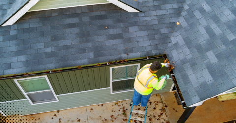 Person cleaning gutter on ladder with clear sky.